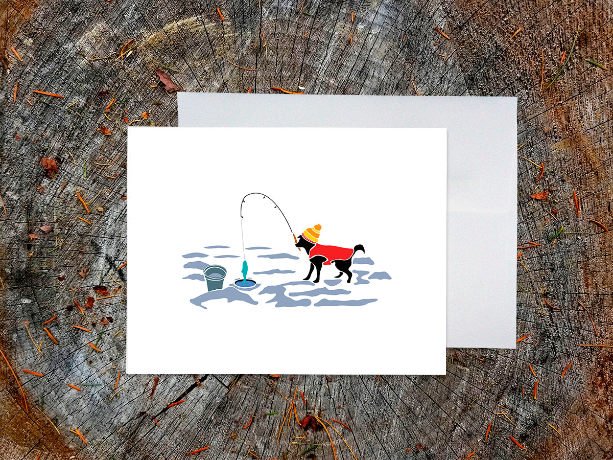 Greeting card with image of a dog with a winter hat and sweater ice fishing with a white envelope in the background and placed on a wooden surface.
