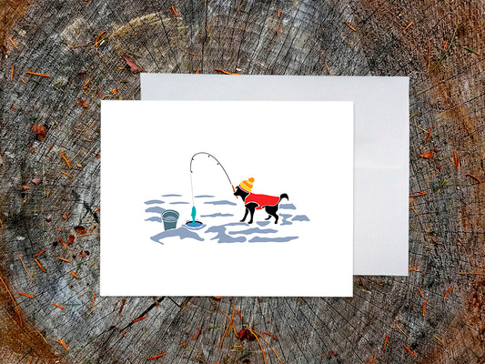 Greeting card with image of a dog with a winter hat and sweater ice fishing with a white envelope in the background and placed on a wooden surface.