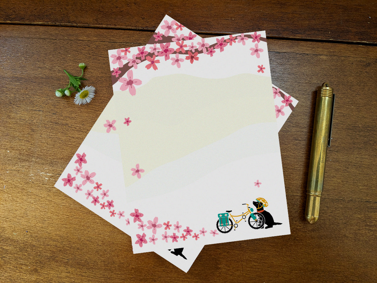 Notepad pages with a black dog next to bicycle among cherry blossoms on wooden background with gold pen.
