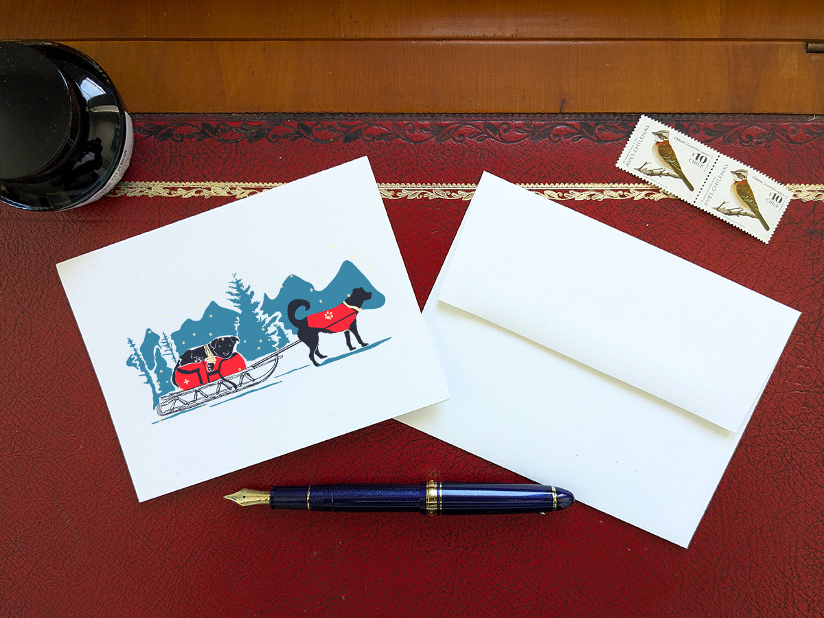 Greeting card with dog in a red jacket pulling a sled with another dog in a wintry landscape on a red desk, next to a blue pen, envelope and stamps.