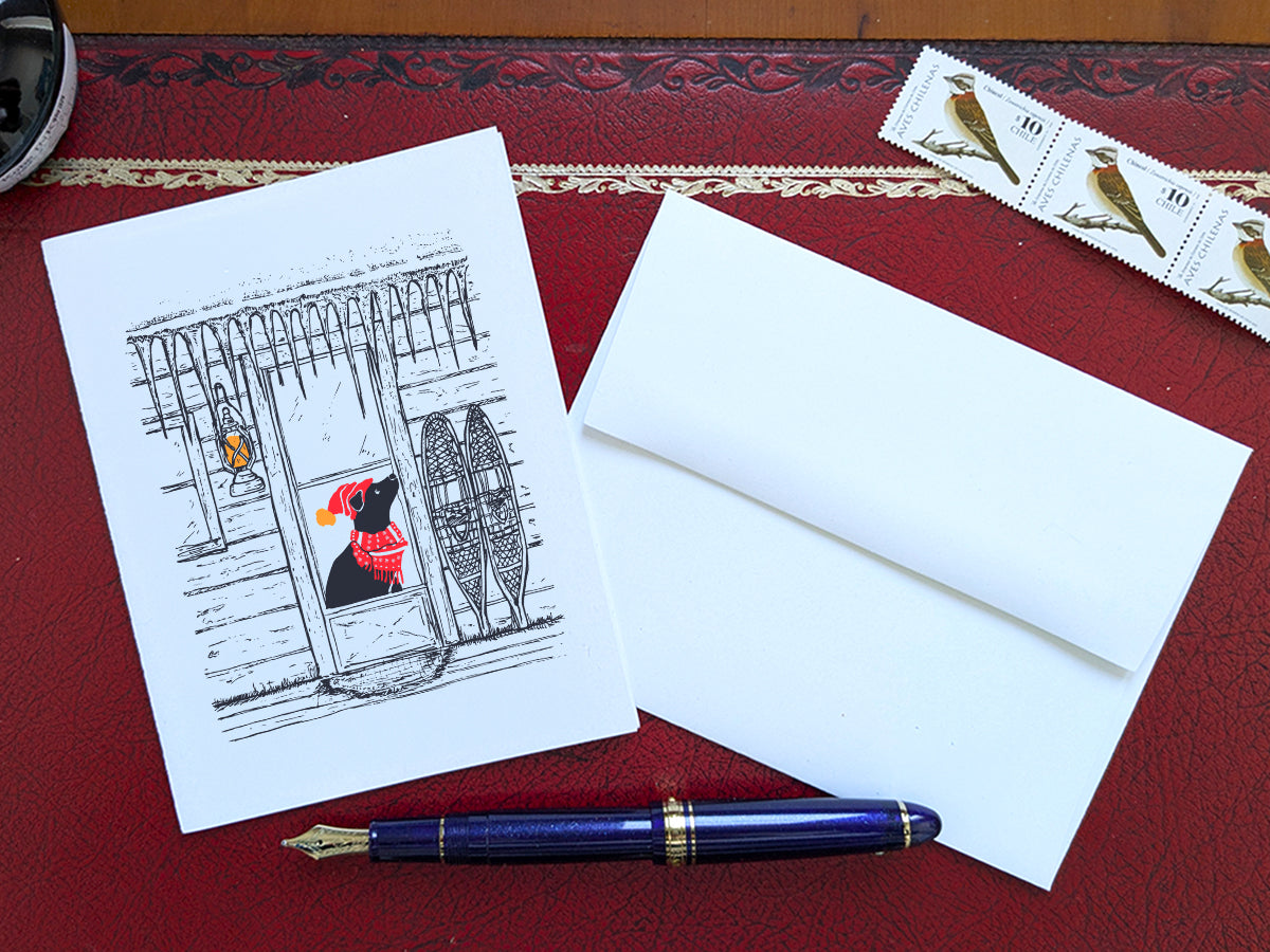 Greeting card with dog with red scarf and hat in a cabin covered in icicles, on a red desk, next to a blue pen, envelope and stamps.