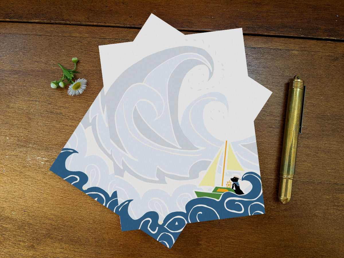 Notepad with illustration of dog sailing big waves on a wood surface and gold pen.