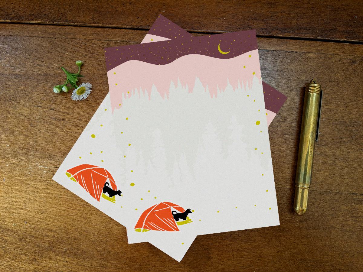 Notepad pages with a black dog in red tent and night sky on wooden background with gold pen.