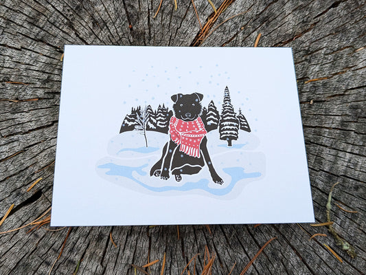 Greeting card with image of black dog with red scarf among a snowy winter landscape.