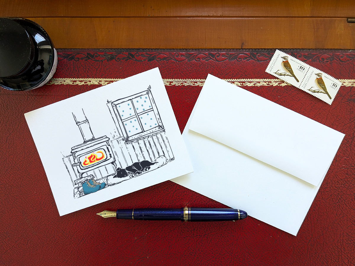 A greeting card with a illustrated scene featuring a wood-burning stove, snow covered window, sleeping dog, and hiking boots, displayed on a red desk with an envelope, blue fountain pen and stamps.