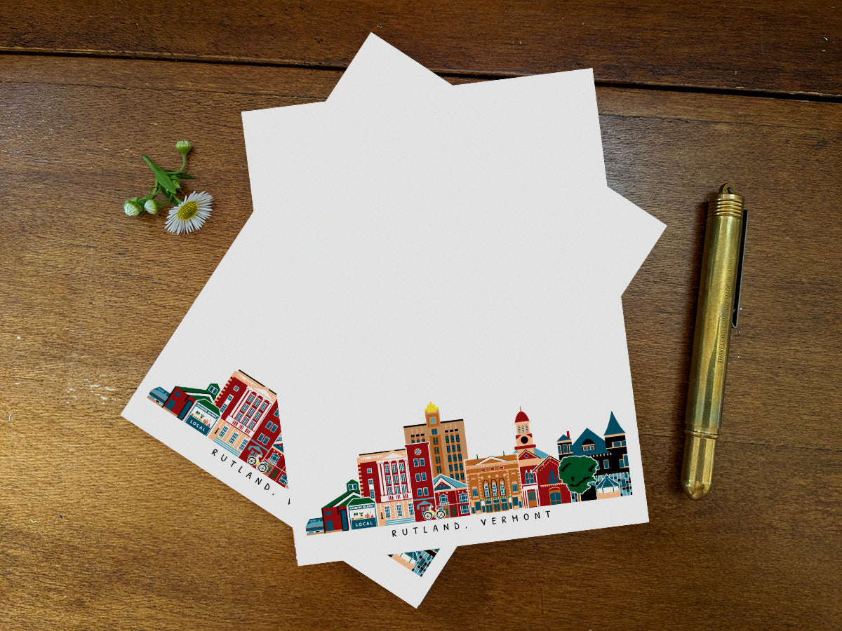 Notepad pages with illustration of Rutland, Vermont buildings in skyline on a wood surface with gold pen