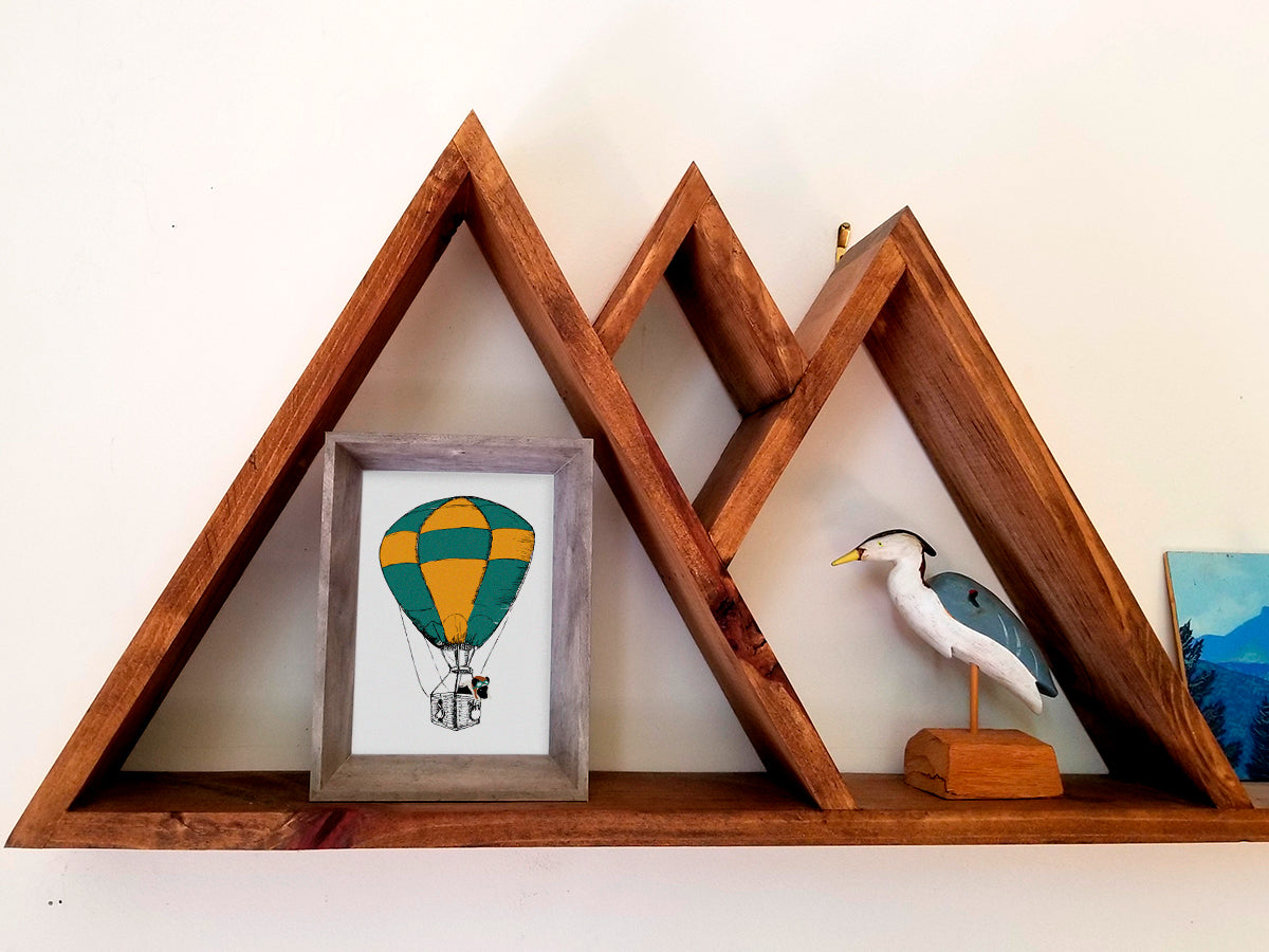 Framed illustration with a green and yellow hot air balloon with a black dog inside the basket on a wooden shelf.