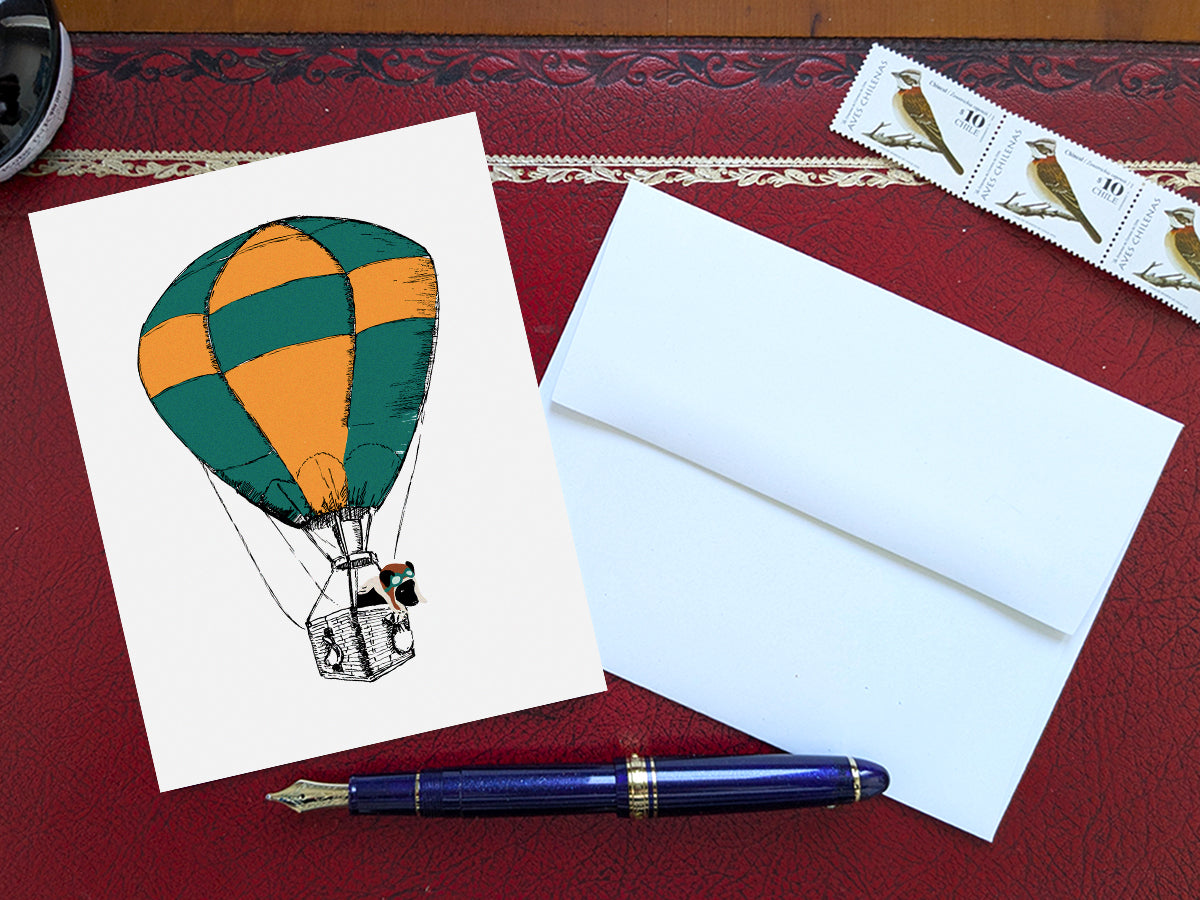 Greeting card with illustration of green and yellow hot air balloon with a black dog inside the basket on a red table with envelope and blue pen and stamps.