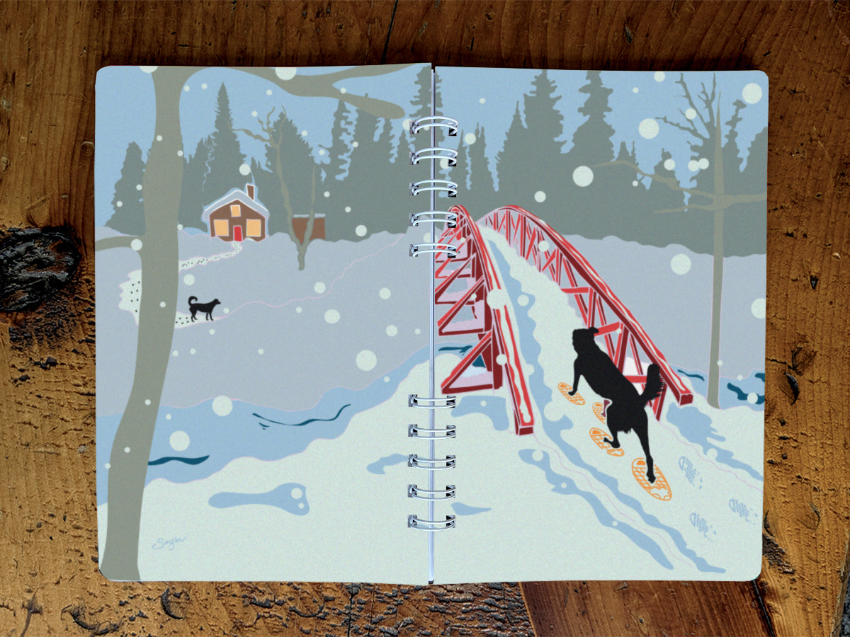A spiral-bound notebook with an illustration of a dog snowshoeing over a bridge toward a cabin and another dog, shown on a wood surface.