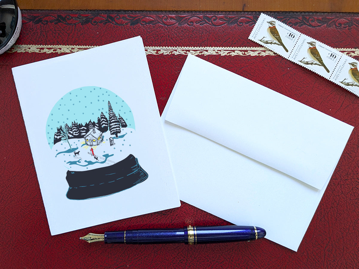 A greeting card with a snow globe illustration with a cabin, skier, and two dogs among a snowy landscape, displayed on a red desk with an envelope, stamps and blue fountain pen.