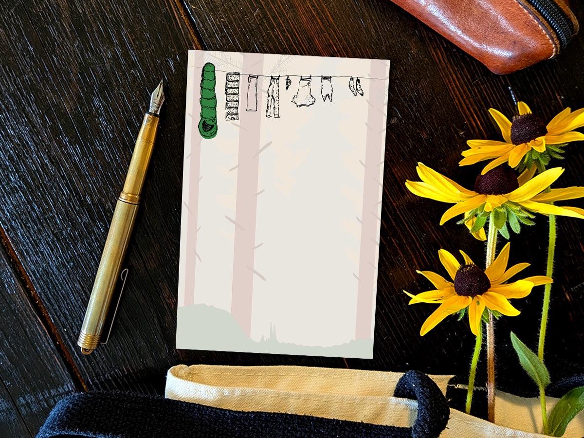 Notepad with illustration of clothes and sleeping bag hanging on a line in between pine trees in a forest, placed on wooden surface with a gold pen, and yellow flowers.