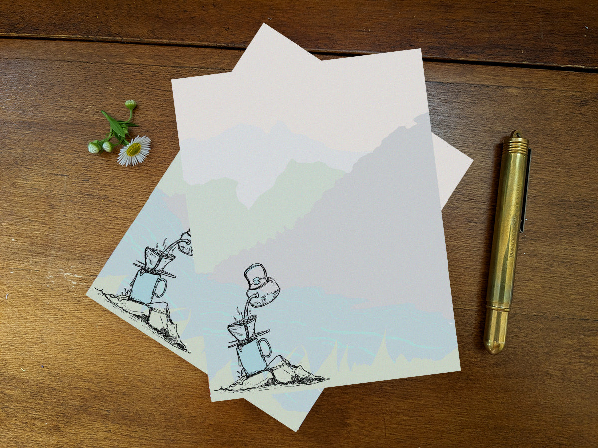 Notepad with illustration of pour-over coffee on a riverbank on a wood surface and gold pen.