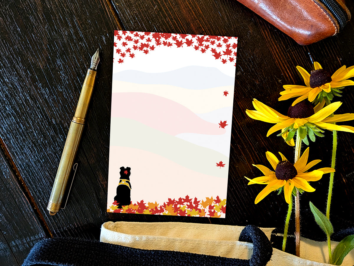 Notepad with an illustration of a black dog sitting among red, orange and yellow fall foliage with colorful rolling hills in the background, placed on a wooden surface with a gold pen, and yellow flowers.