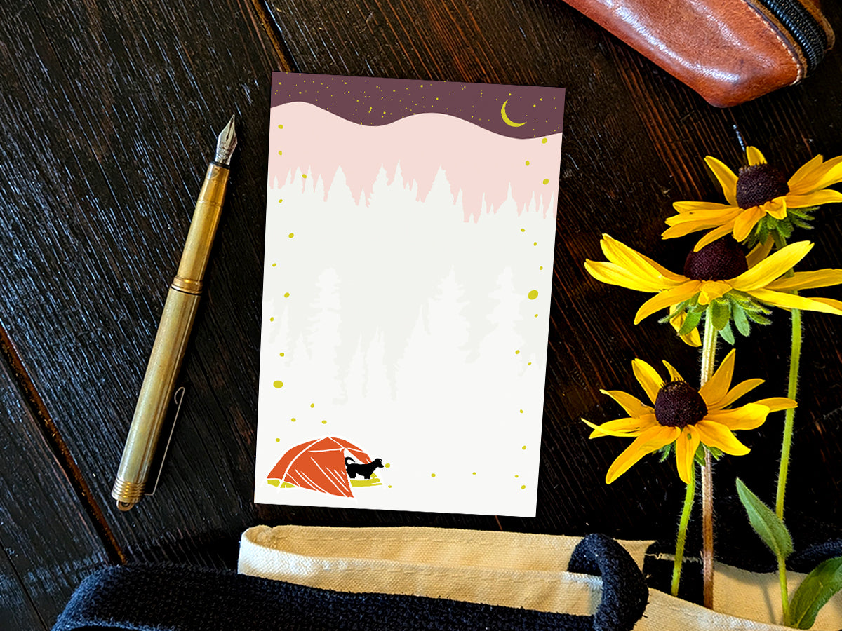 Notepad with an illustration of a black dog in red tent camping under night sky with fireflies, placed on a wood surface with a gold pen, and yellow flowers.