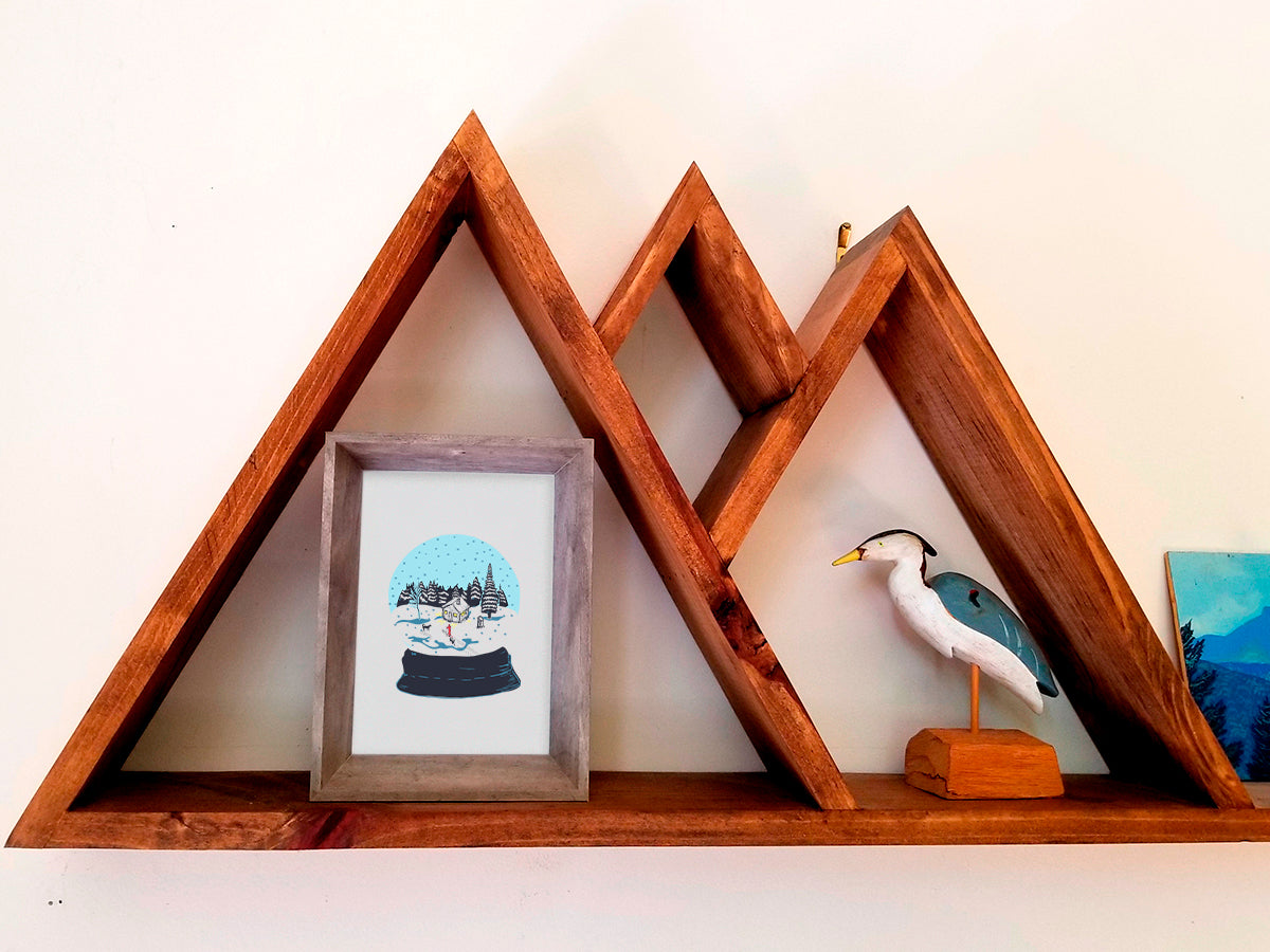 A framed art print featuring a snow globe illustration with a cabin, skier, and two dogs among a snowy landscape, displayed on a wooden shelf.