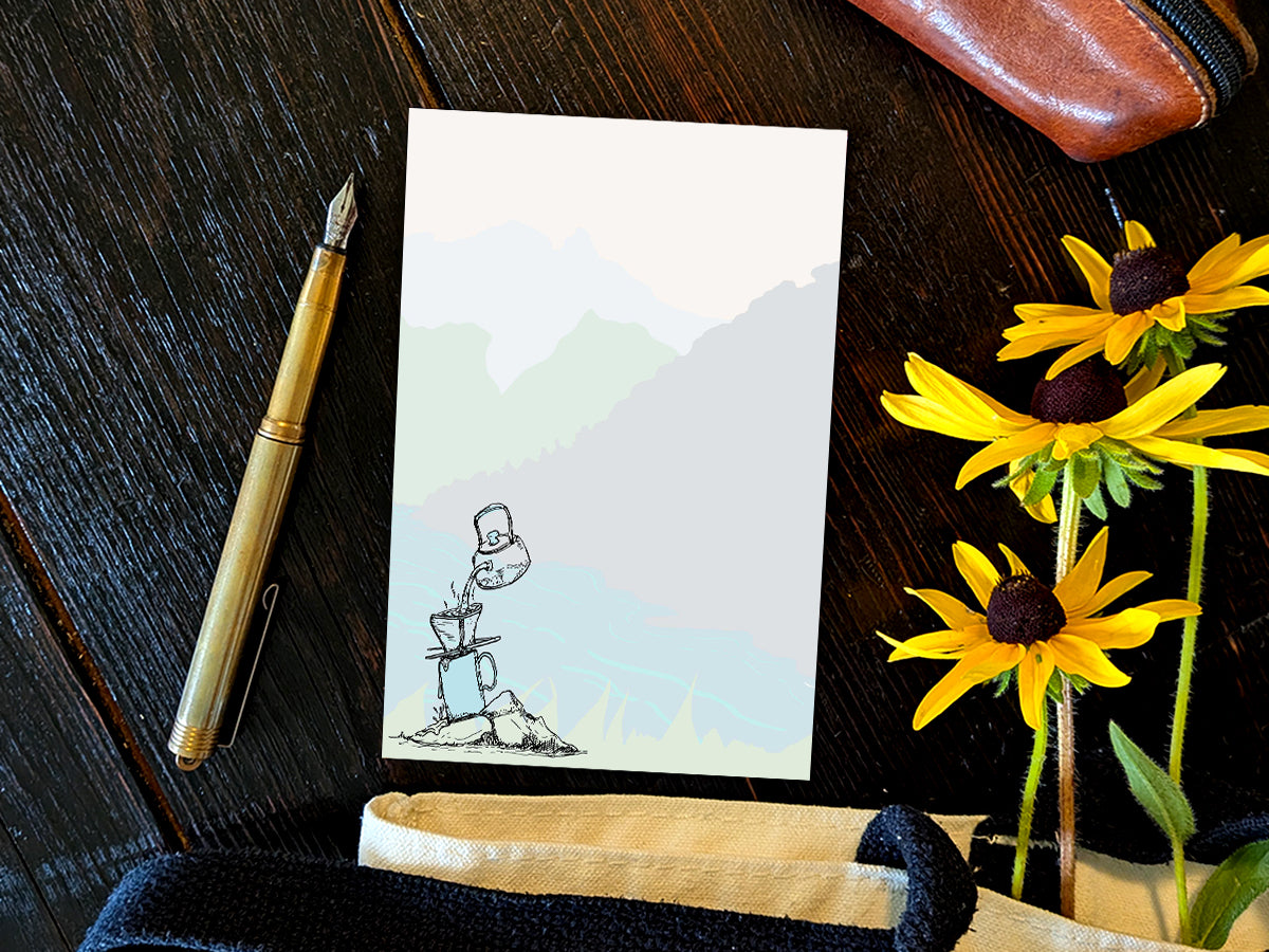 Notepad with illustration of pour-over coffee on a riverbank, placed on a wood surface with a gold pen, and yellow flowers.