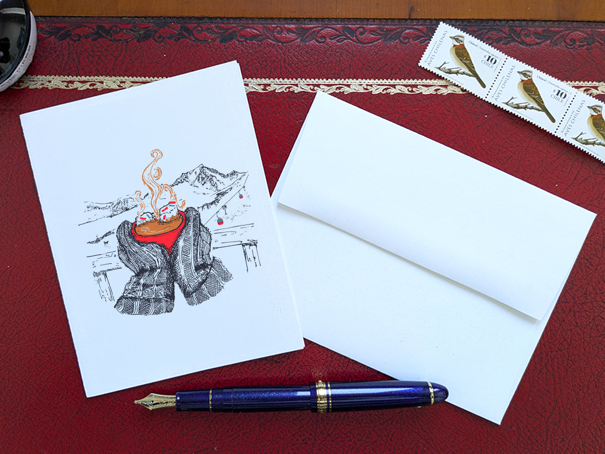 A greeting card with an illustration of a red, steaming cup of hot chocolate being held by big mittens, with mountains in the background, on a red desk next to a white envelope, blue fountain pen and stamps.