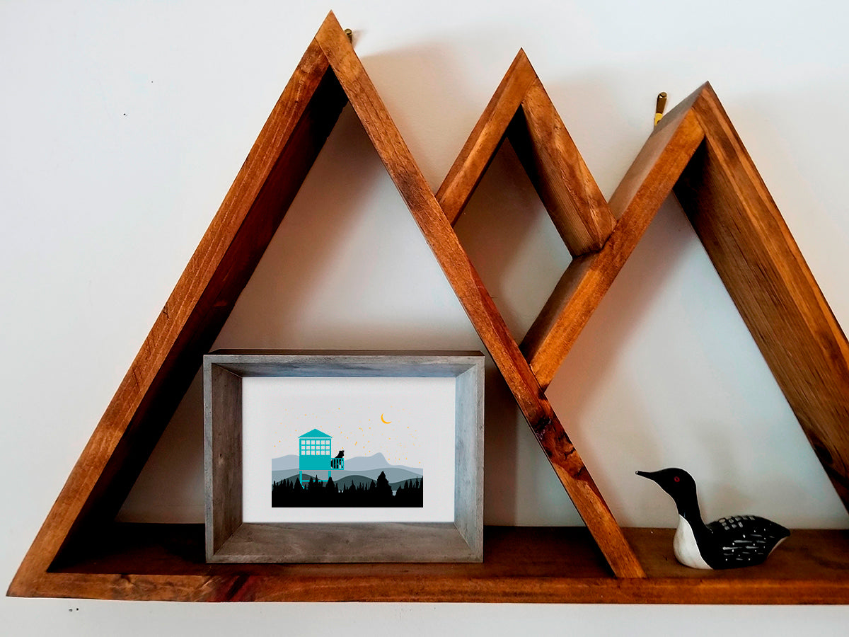 Framed illustration of a dog in a fire tower overlooking the mountains on a wooden shelf.