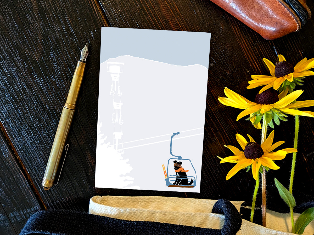 Notepad with illustration of a dog with goggles and orange scarf riding a ski lift, placed on a wooden surface with a gold pen, and yellow flowers.
