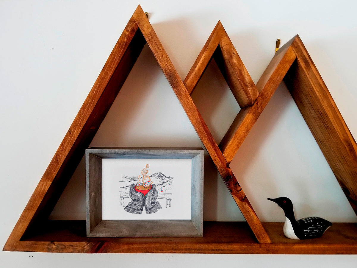 Framed art print illustration of thick winter mittens holding a red cup of hot chocolate, with mountains and chair lift in the background, displayed on a wooden shelf.