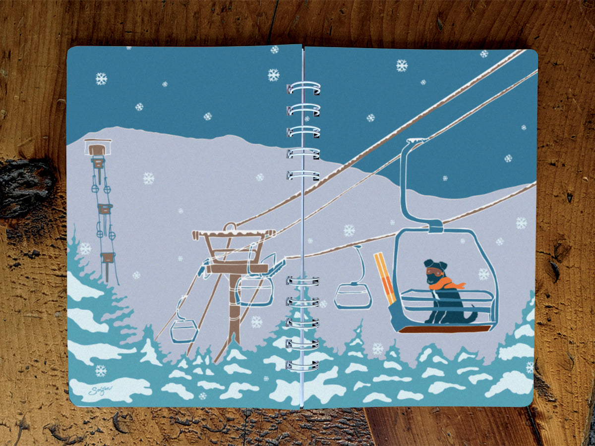A spiral-bound notebook with an illustration of a dog riding a ski lift, on a wood surface.