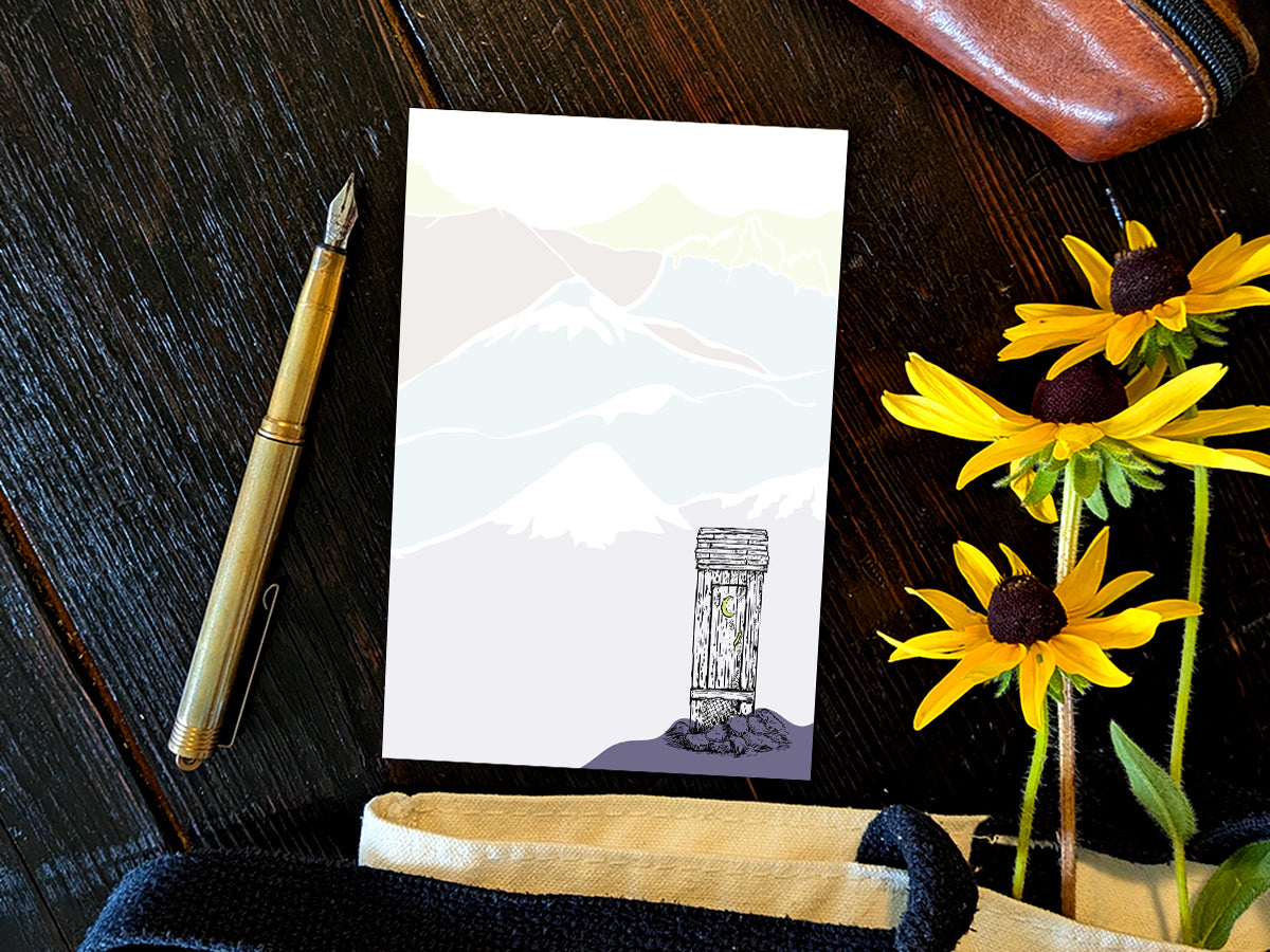 Notepad with an illustration of an outhouse in the mountains, placed on a wood surface with a gold pen, and yellow flowers.