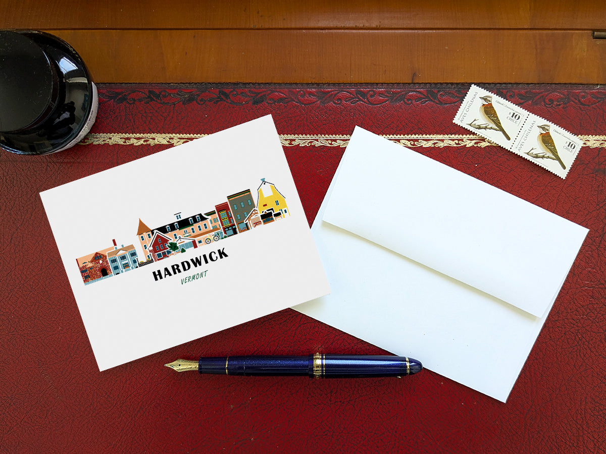 Greeting card with image of buildings arranged in a city skyline with the text Hardwick Vermont on a red table with blue pen, stamps and envelope.
