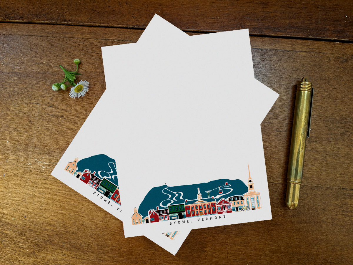 Notepad pages with illustration of Stowe, Vermont buildings in skyline on a wood surface with gold pen