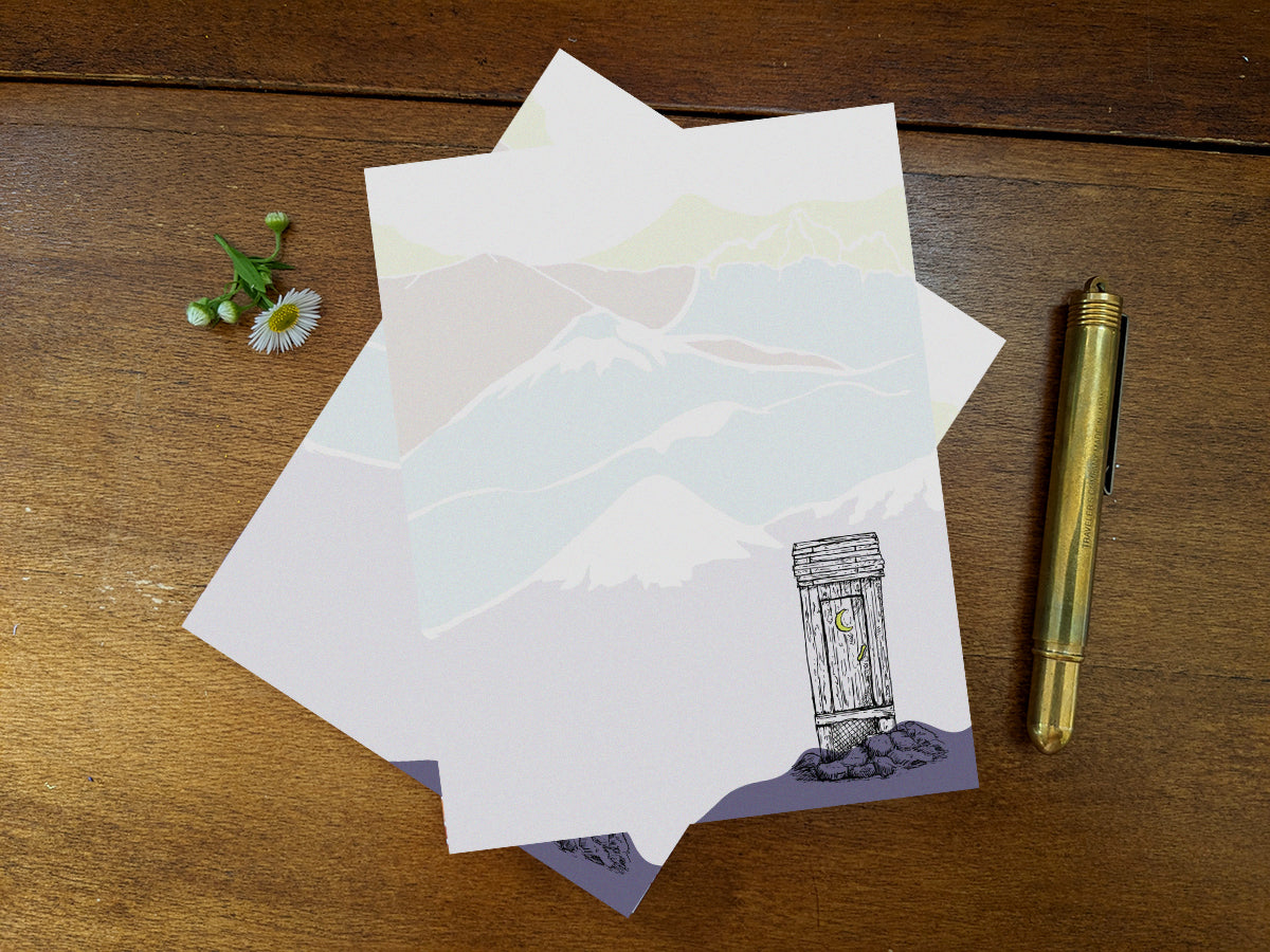 Notepad with an illustration of an outhouse in the mountains on a wood surface and gold pen.