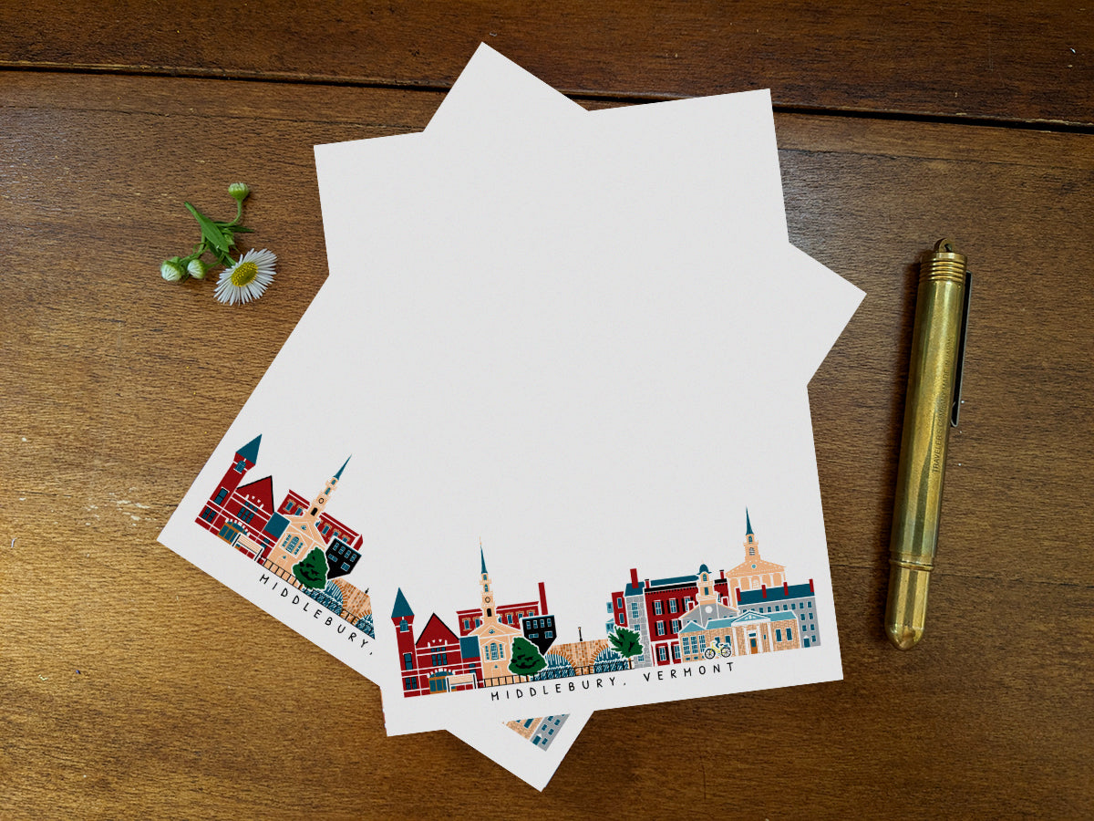 Notepad pages with illustration of Middlebury, Vermont buildings in skyline on a wood surface with gold pen