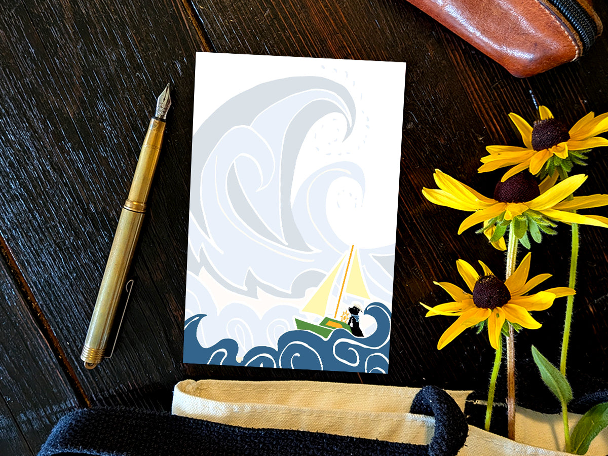 Notepad with illustration of dog sailing big waves on a wood surface with a gold pen, and yellow flowers.