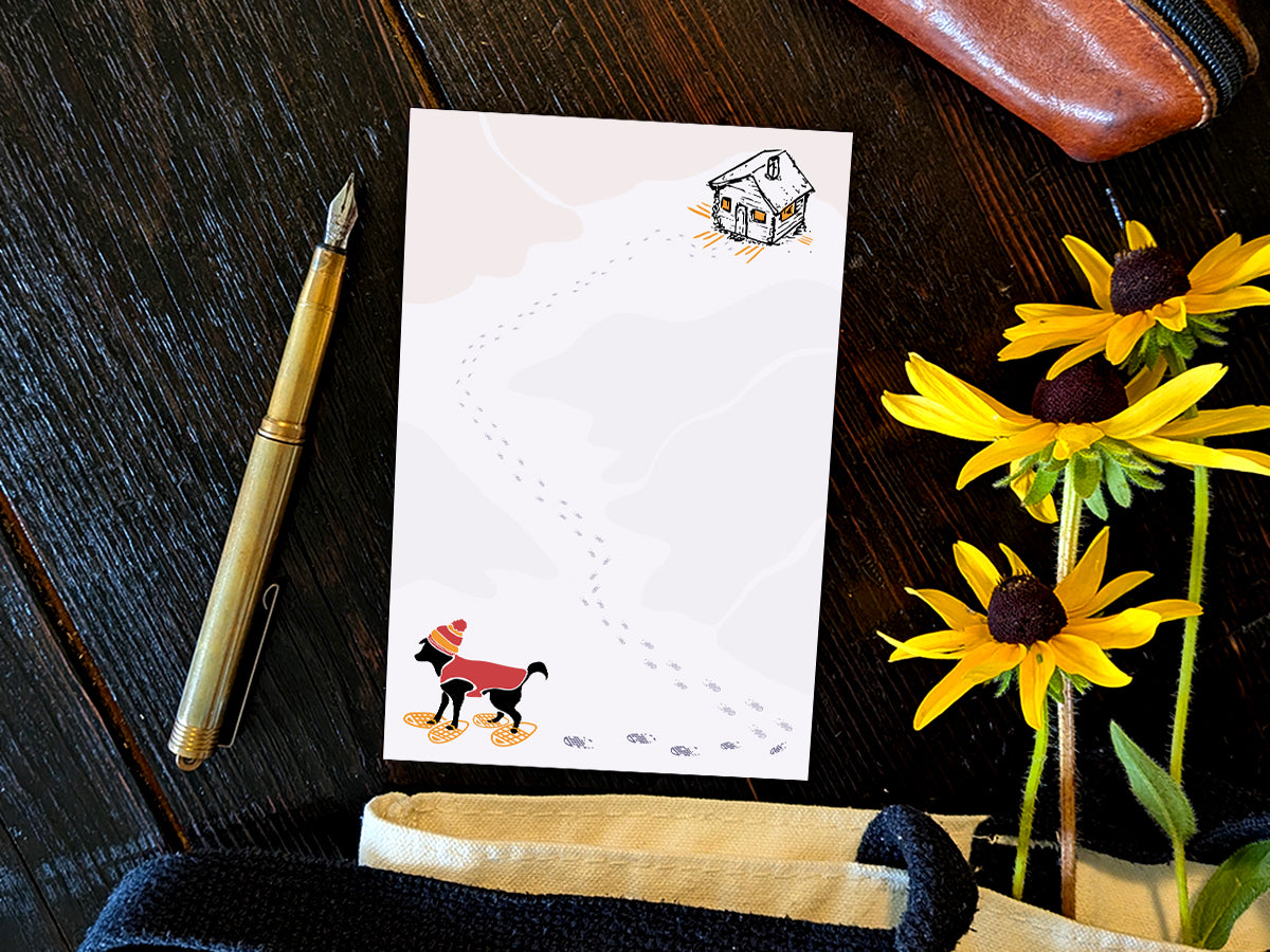 Notepad with illustration of a black dog with winter hat, red jacket, and snowshoes, with a trail a footprints leading from a rustic cabin in the background, placed on a wooden surface with a gold pen, and yellow flowers.
