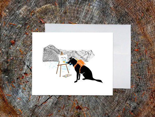 Greeting card with illustration of black dog painting in the mountains, with a white envelope placed on a wooden surface.