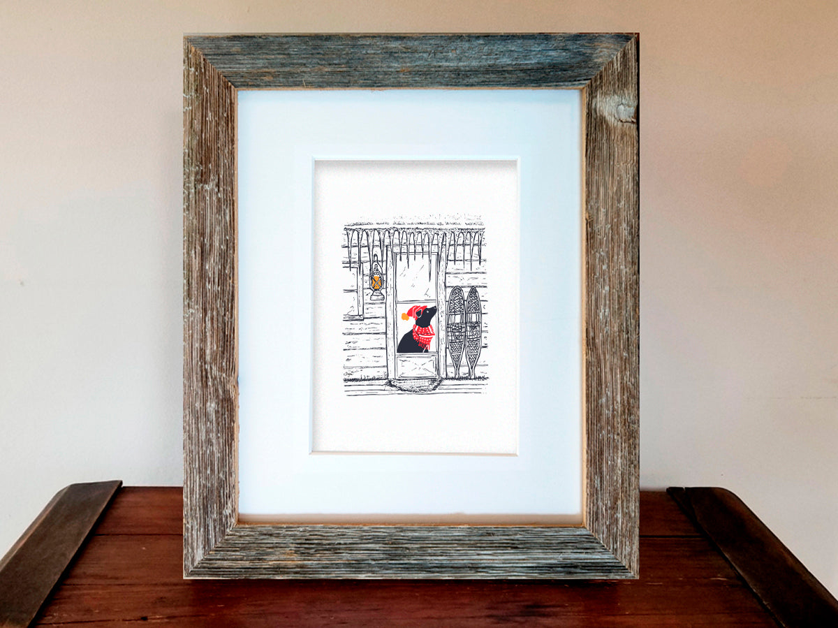 Framed art print featuring an illustration of a dog with red scarf and hat in a cabin covered in icicles, displayed on a wooden shelf.