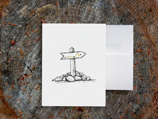 White card with a black ink drawing of a trail marker on a wooden surface.
