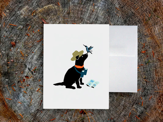 White card with drawing of a dog with binoculars and a hummingbird on a wooden surface.