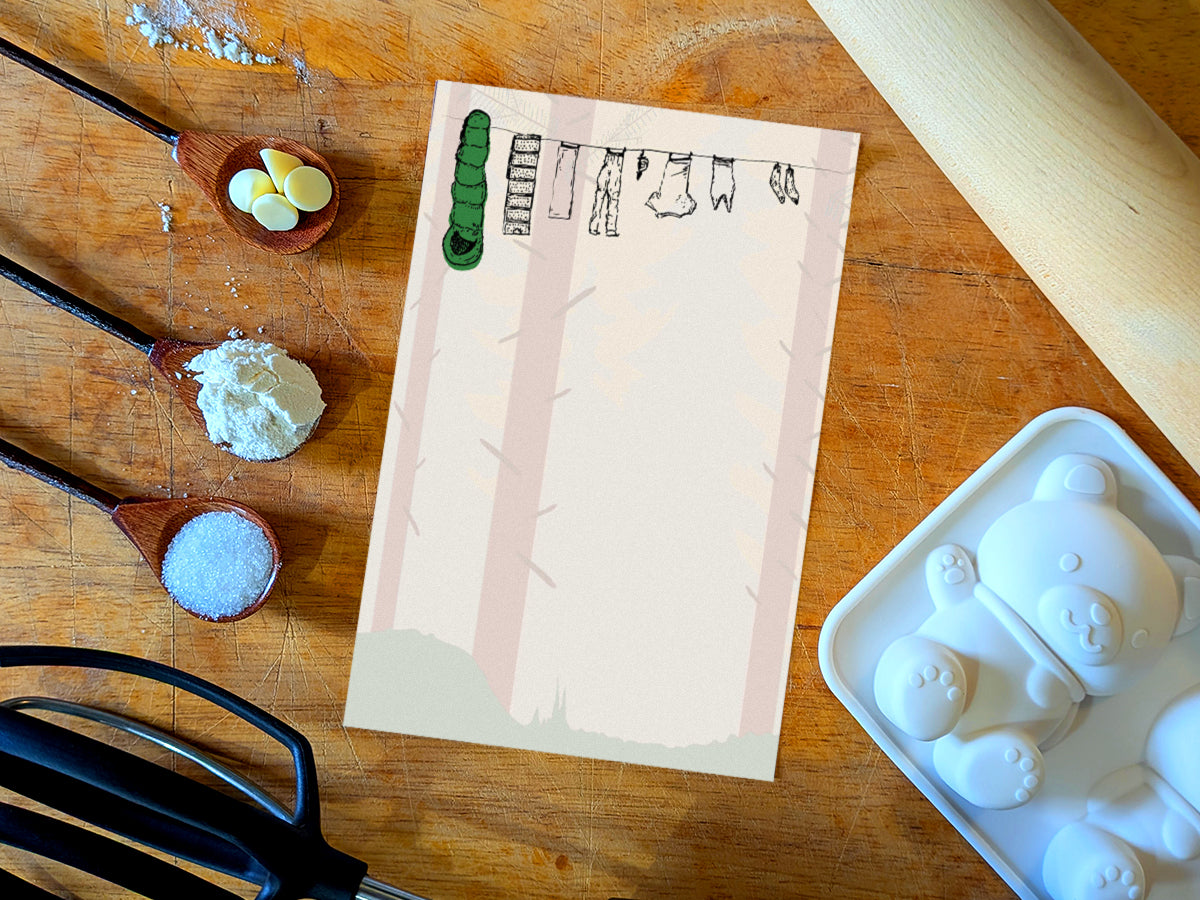 Notepad with illustration of clothes and sleeping bag hanging on a line in between pine trees in a forest, placed on wooden surface with baking supplies.