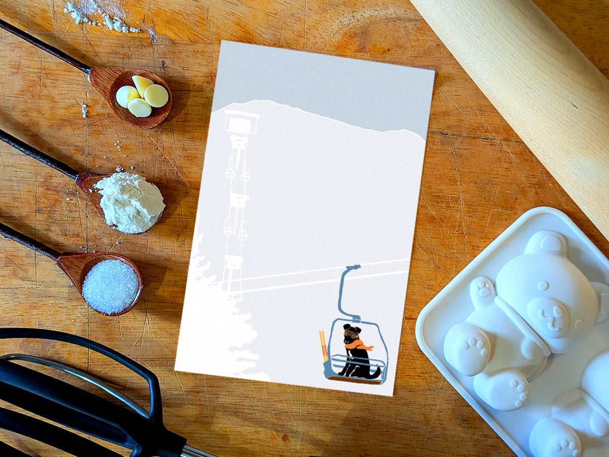 Notepad with illustration of a dog with goggles and orange scarf riding a ski lift, placed on a wooden surface with baking supplies.