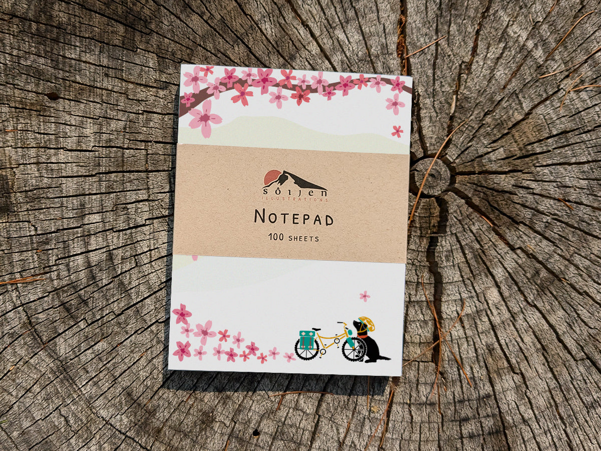 Notepad with a black dog next to bicycle among cherry blossoms on wooden background.