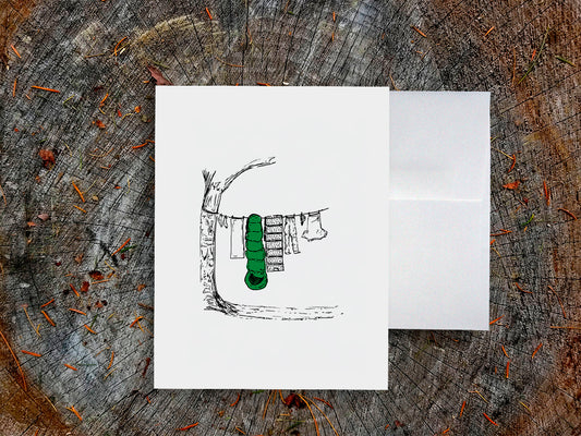 White card with a black ink drawing of laundry hanging from a tree on a wooden surface.