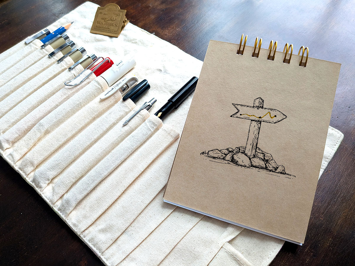 Notebook with a drawing of a trail marker, next a pen case on a wood surface.