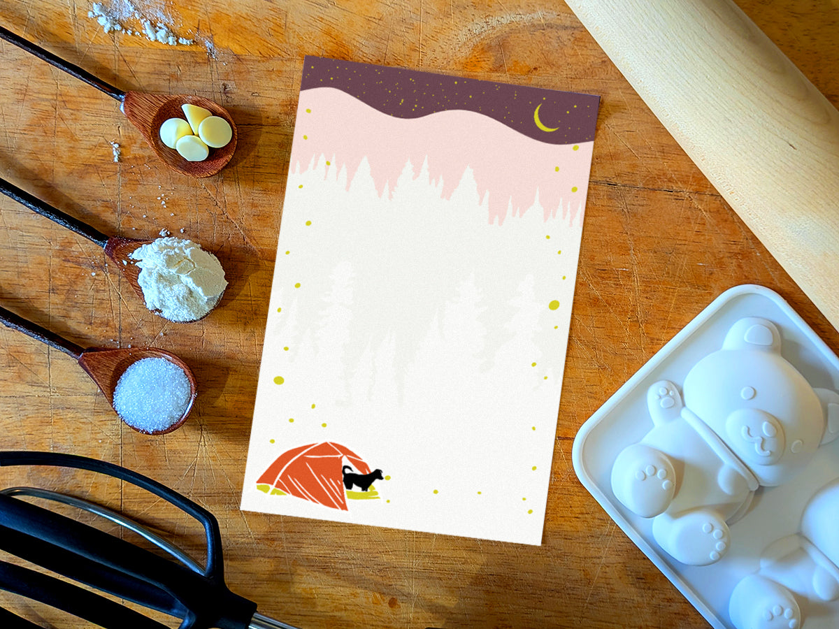 Notepad with an illustration of a black dog in red tent camping under night sky with fireflies, placed on a wood surface with baking supplies.