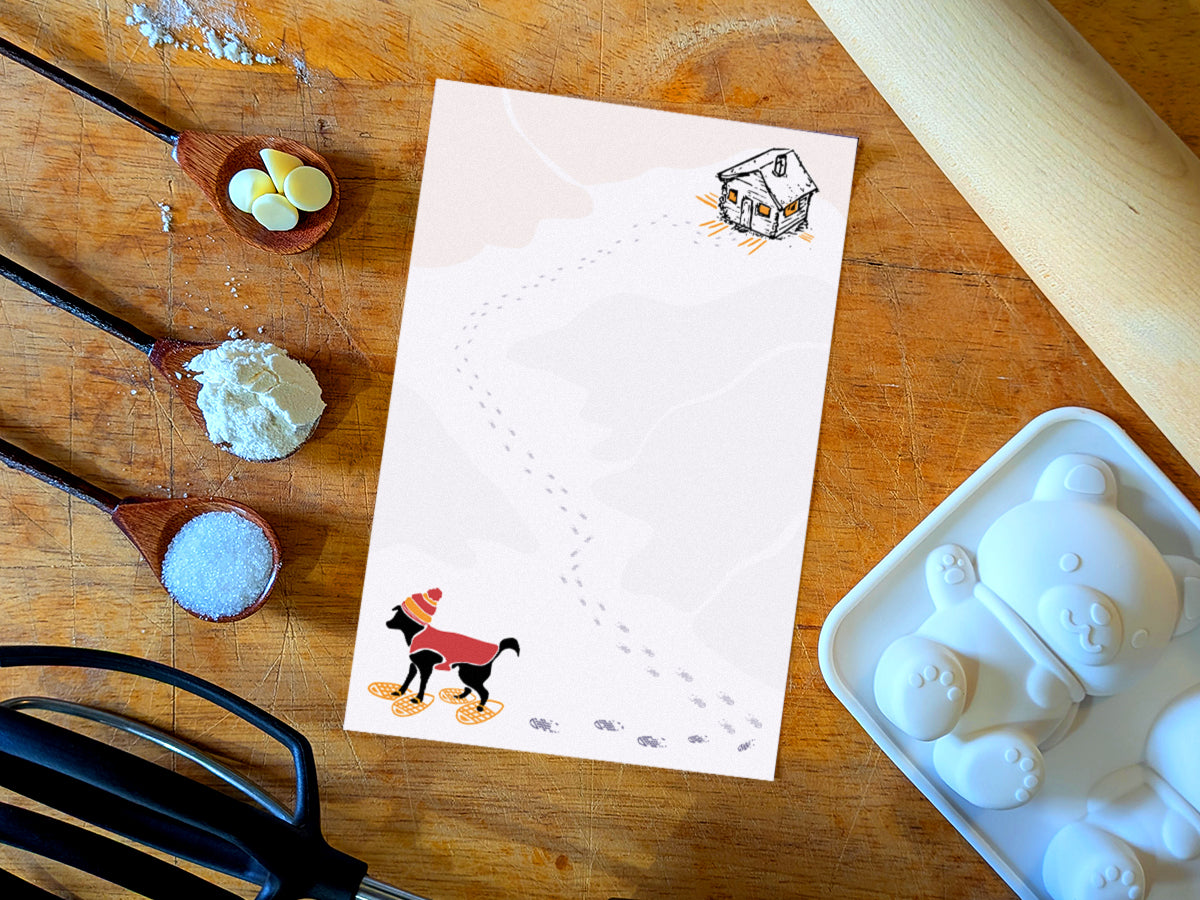 Notepad with illustration of a black dog with winter hat, red jacket, and snowshoes, with a trail a footprints leading from a rustic cabin in the background, placed on a wooden surface with baking supplies.