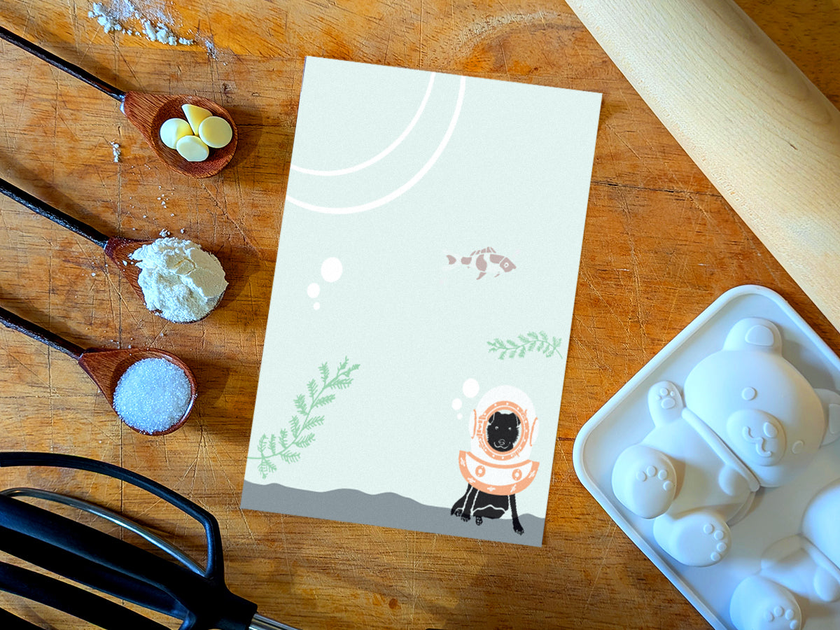 Notepad with a dog underwater with old school diving helmet and red fish, placed on a wood surface with baking supplies.