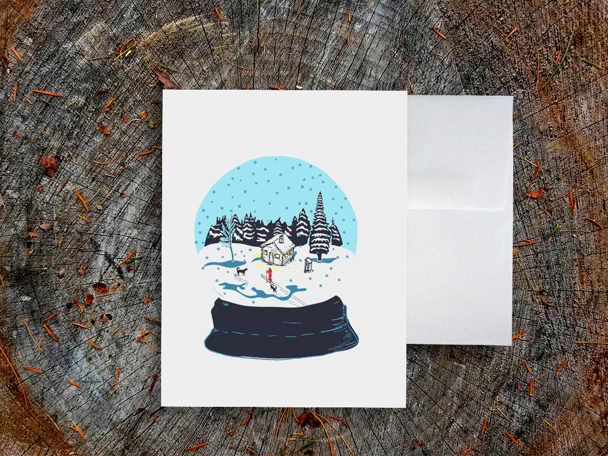 A greeting card with a snow globe illustration with a cabin, skier, and two dogs among a snowy landscape, displayed on a wooden surface with an envelope.