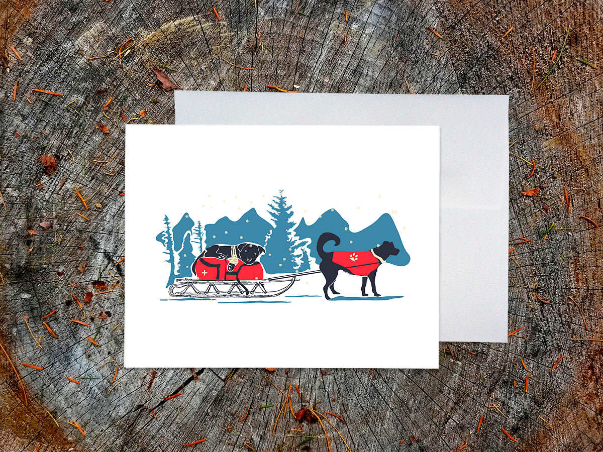 Greeting card with dog in a red jacket pulling a sled with another dog in a wintry landscape, on a wooden surface with white envelope.
