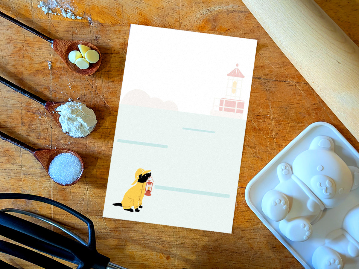 Notepad with an illustration of a dog in yellow hat and jacket holding a red lantern with a lighthouse in the background, placed on a wood surface with baking supplies.