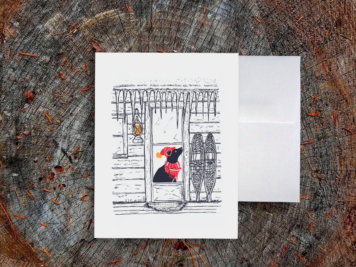 Greeting card with dog with red scarf and hat in a cabin covered in icicles, on a wooden surface with white envelope.