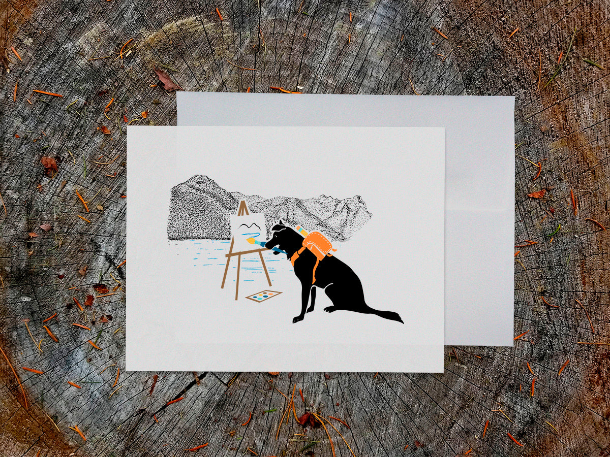 White card with drawing of dog painting in the mountains on a wooden surface.