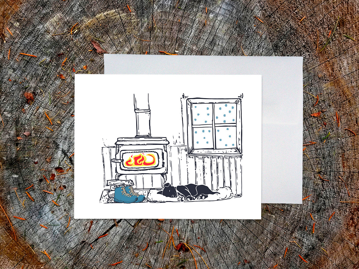A greeting card with a illustrated scene featuring a wood-burning stove, snow covered window, sleeping dog, and hiking boots, displayed on a wooden background with an envelope behind it.